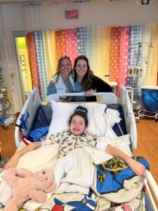 Photograph of a young girl named Collyns in lying in a hospital bed posing with friends.