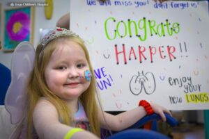 Photograph of a young girl named Harper smiling in front of a white board that says "Congrats Harper" with a drawing of lungs