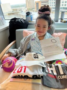 Photograph of a young girl Collyns recovering in a hospital, posing with several HHT branded items and a letter.