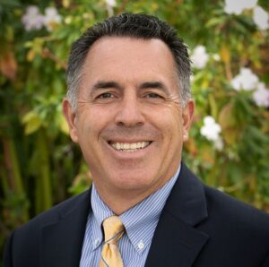 Smiling man in a dark suit and yellow tie outdoors in front of greenery
