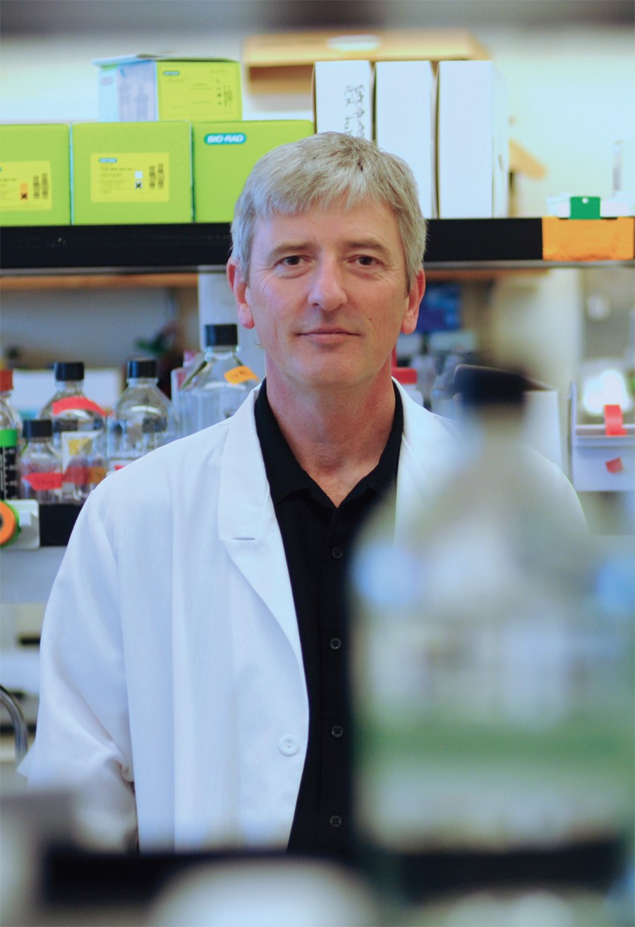 A man in a white lab coat stands among shelves and glassware in a laboratory.
