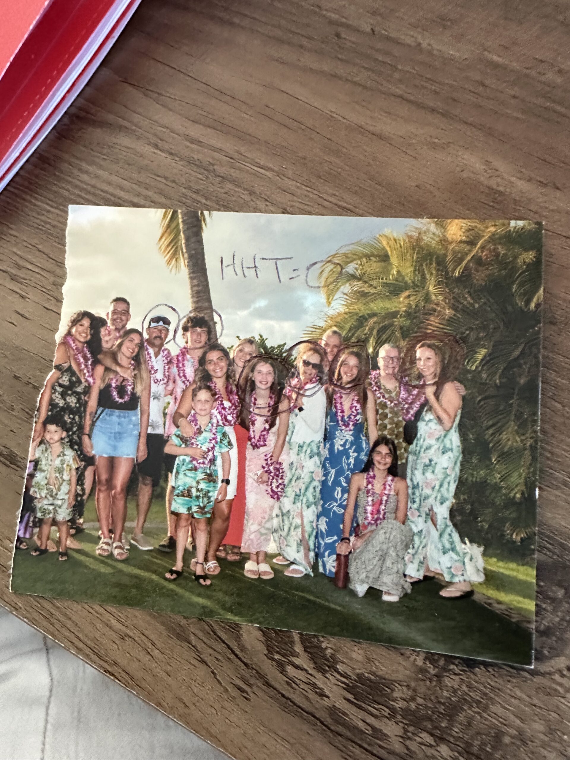 A printed group photo of about fourteen people in floral attire and leis, resting on a wooden table.