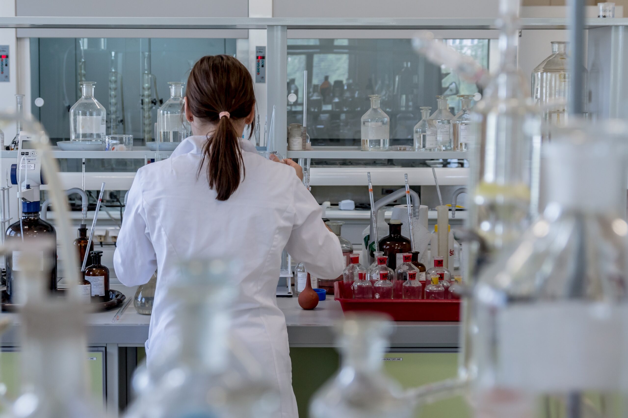 A person in a white lab coat seen from behind, working at a laboratory bench with glassware.