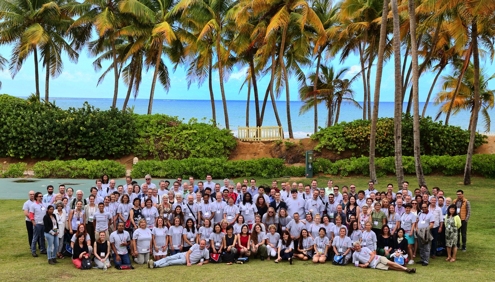 A large group of conference attendees posed on a lawn under palm trees.