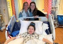 Photograph of a young girl named Collyns in lying in a hospital bed posing with friends.